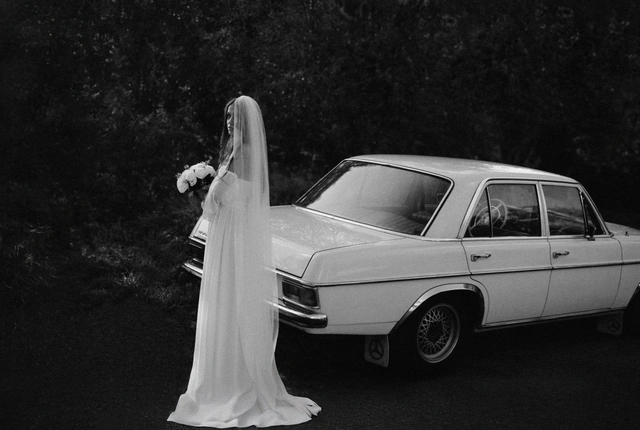 Czarno-biała fotografia panny młodej obok białego samochodu, szeroki kadr